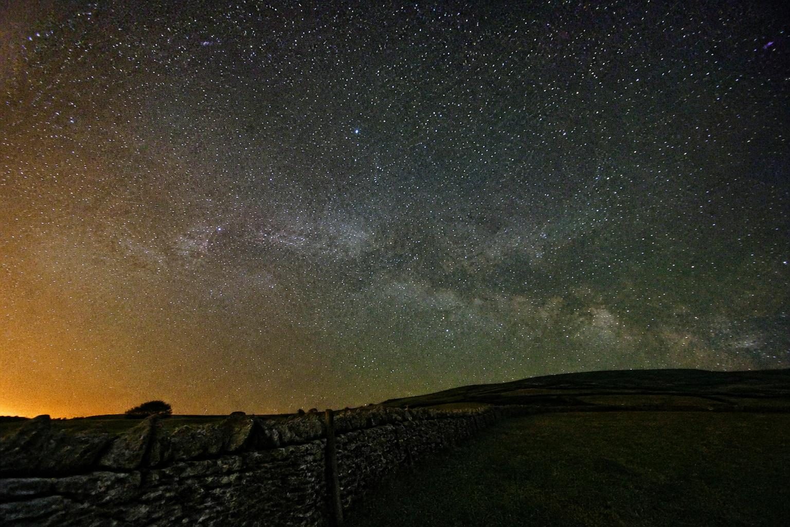 Beginner's first Milky Way: core low over a dry stone wall, orange town glow on the left, very noisy.